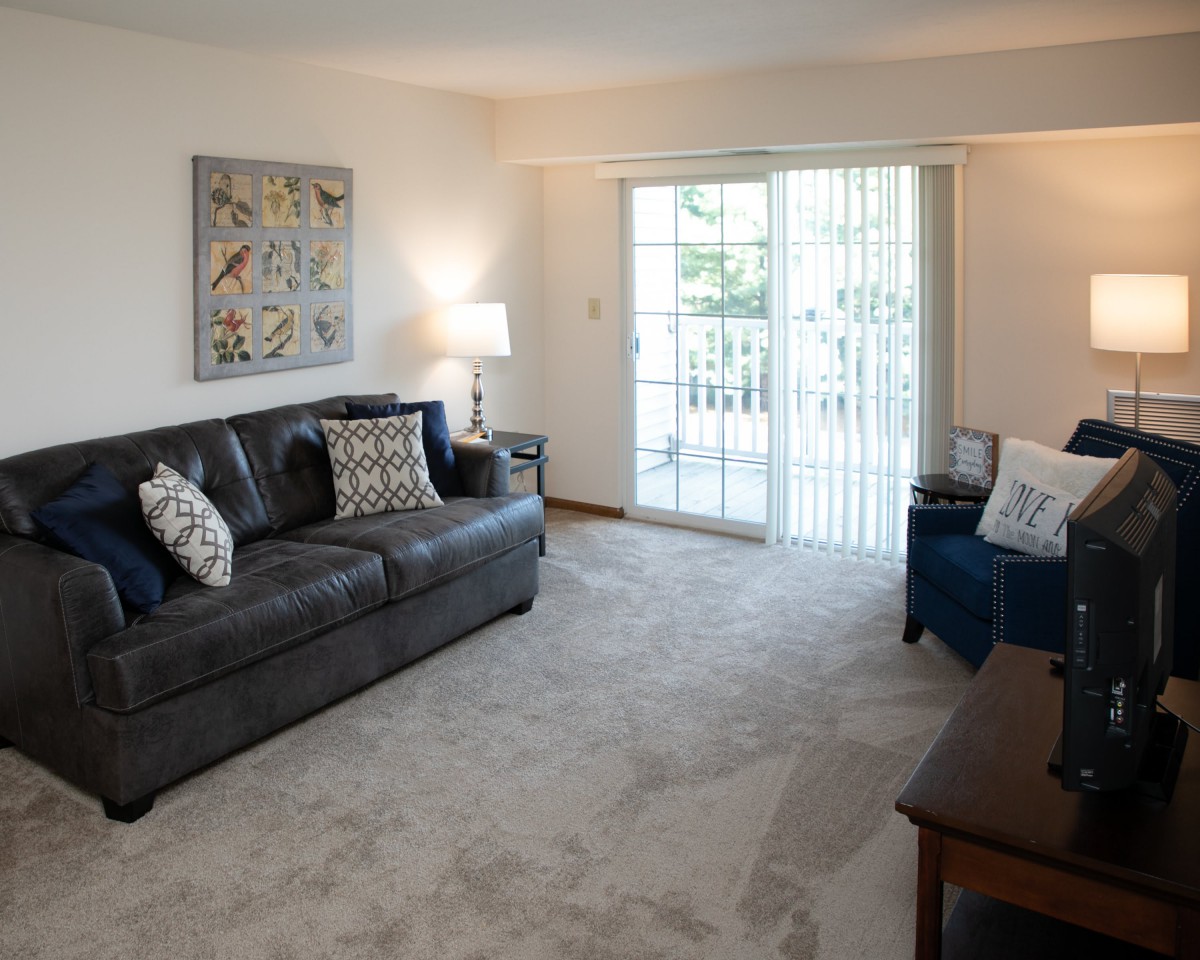 A cozy living room with a dark leather sofa, decorative pillows, and a sliding glass door leading to a balcony.