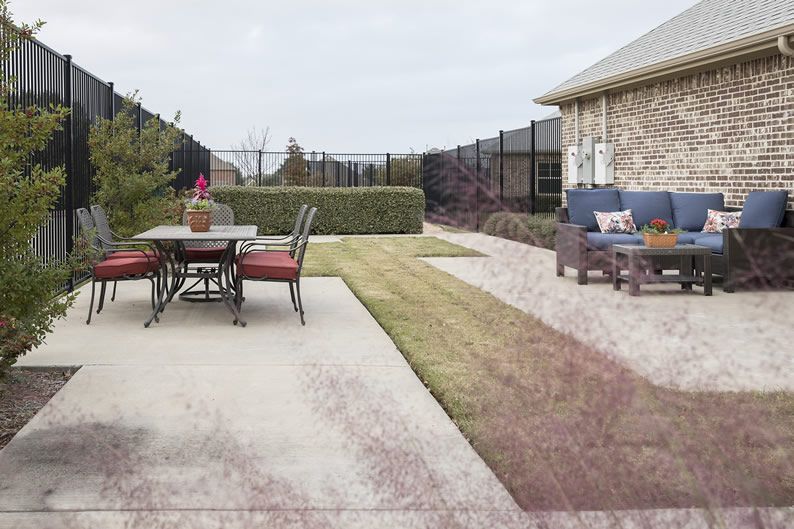 A cozy backyard patio with a table and chairs set on a concrete surface, surrounded by a grassy lawn and bordered by a fence.