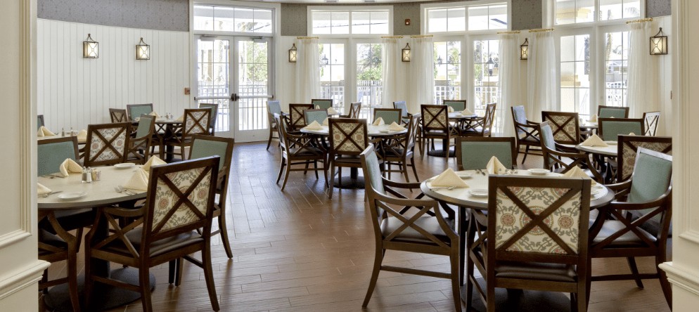 An elegantly set dining room with multiple tables and chairs, well-lit by natural light from large windows.