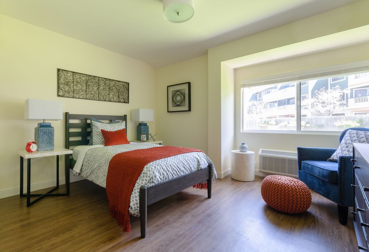 A modern bedroom with a single bed and orange accents, featuring ample natural light from the window.