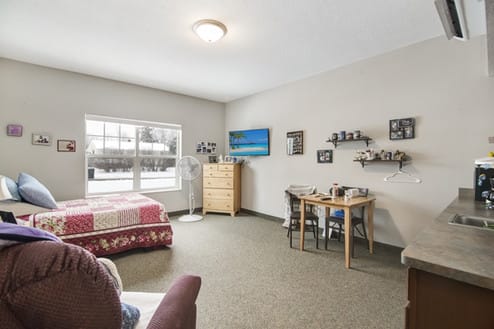 A cozy bedroom with a large window, featuring a bed, dresser, table, and decorative shelves.
