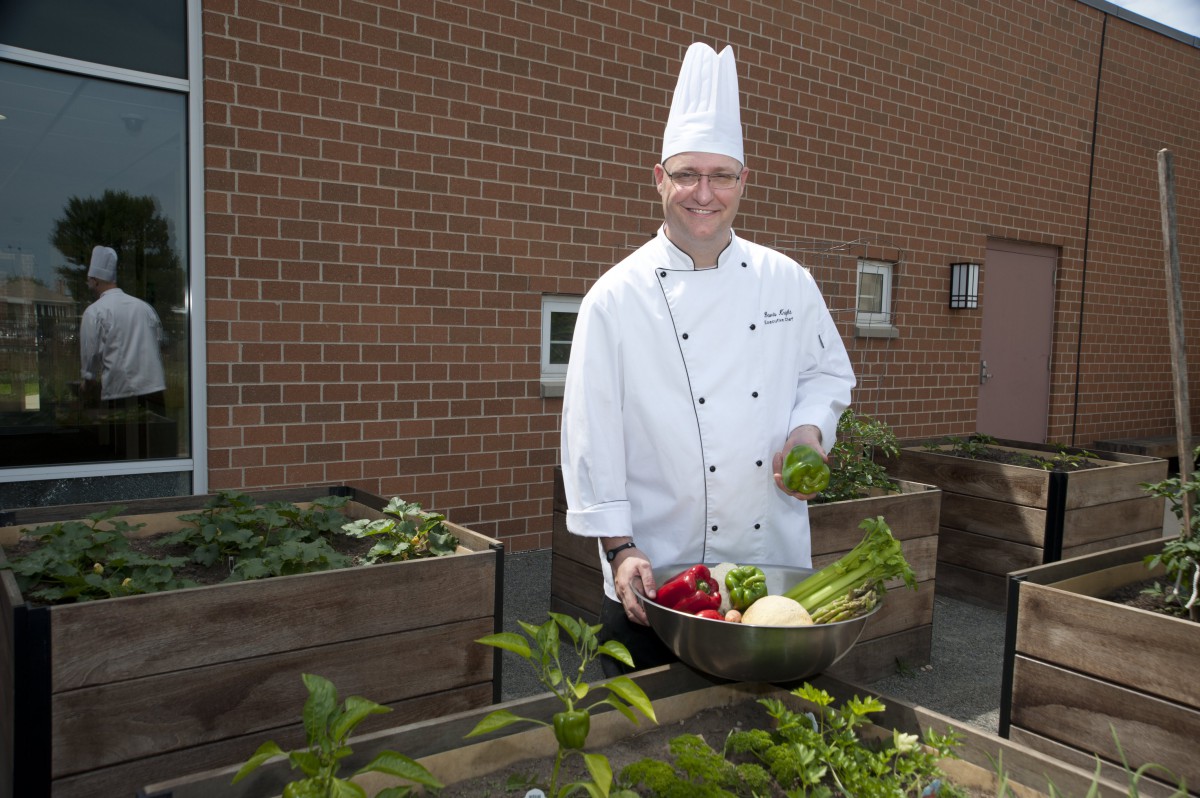 A chef in a white uniform holding fresh vegetables in a garden.