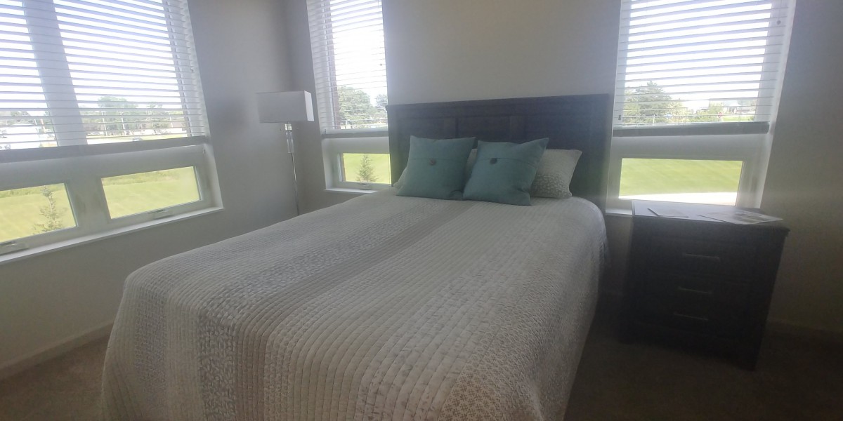 A cozy bedroom with a neatly made bed and decorative pillows, illuminated by natural light from surrounding windows.