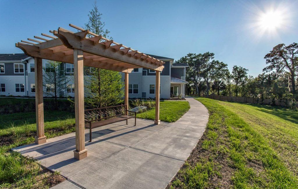 A peaceful outdoor area with a pergola-covered bench, a sidewalk, and a residential building in the background.