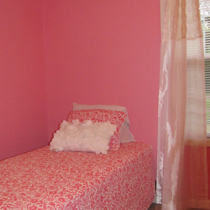 A cozy bedroom with pink walls and a single bed adorned with decorative pillows next to a window.