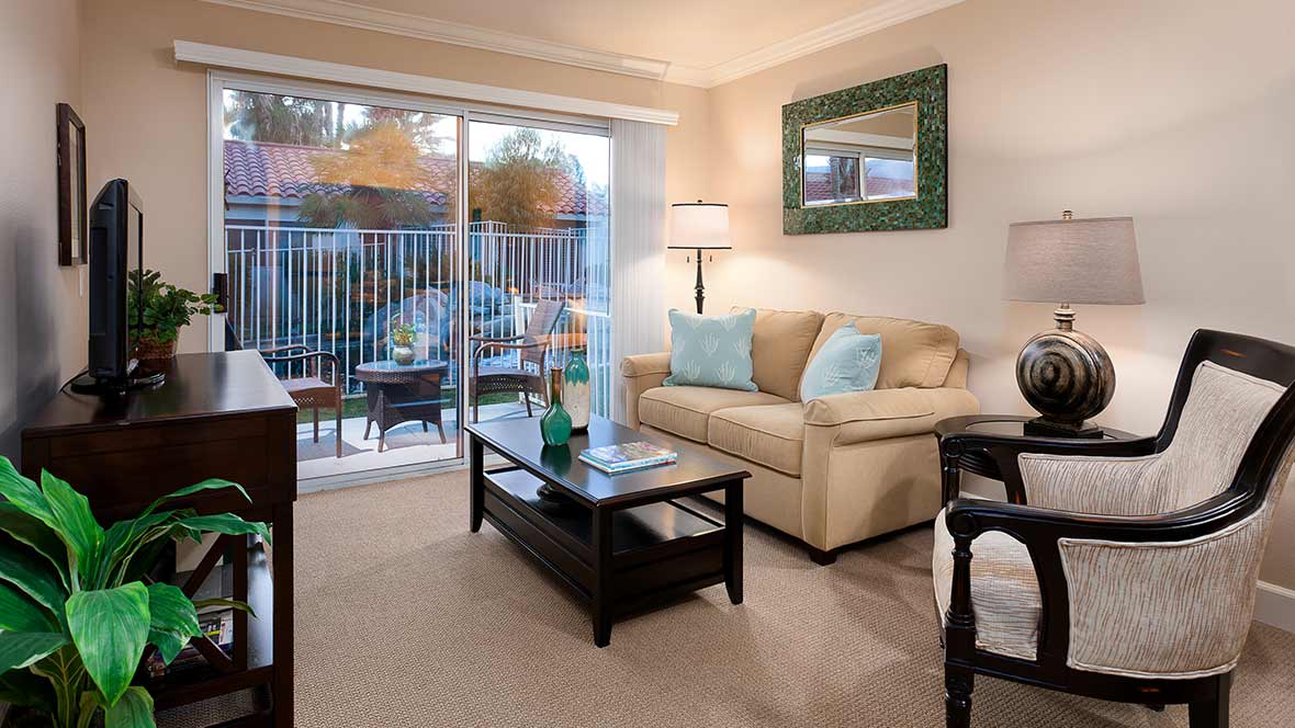 A cozy living room features a beige sofa with blue decorative pillows, a dark wood coffee table, and a large mirror. Natural light streams in through the sliding glass doors leading to an outdoor patio space.