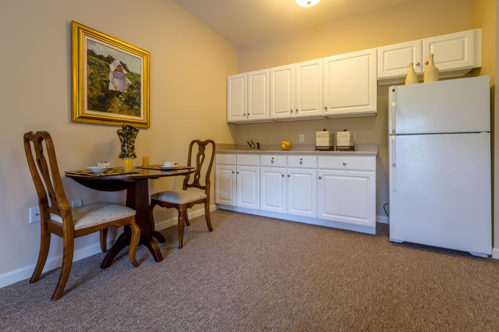 A cozy kitchen and dining area featuring white cabinets, a refrigerator, a small table with two chairs, and carpeted flooring.