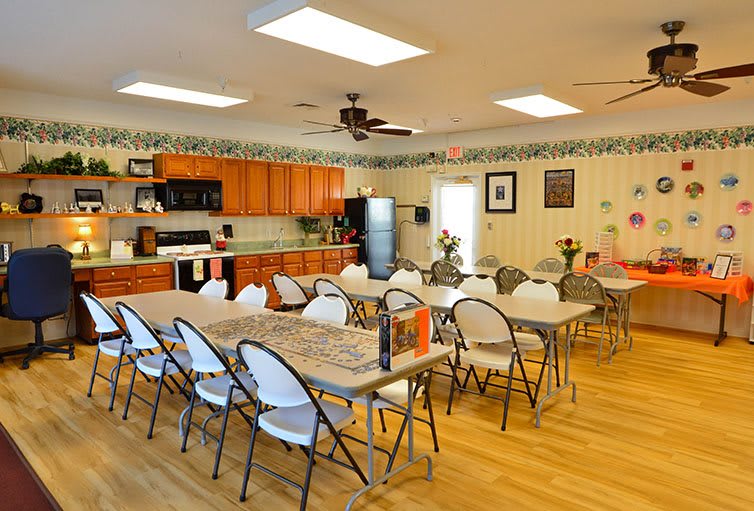 A spacious community meeting room with tables and chairs set up, adjacent to a kitchen with wooden cabinets.