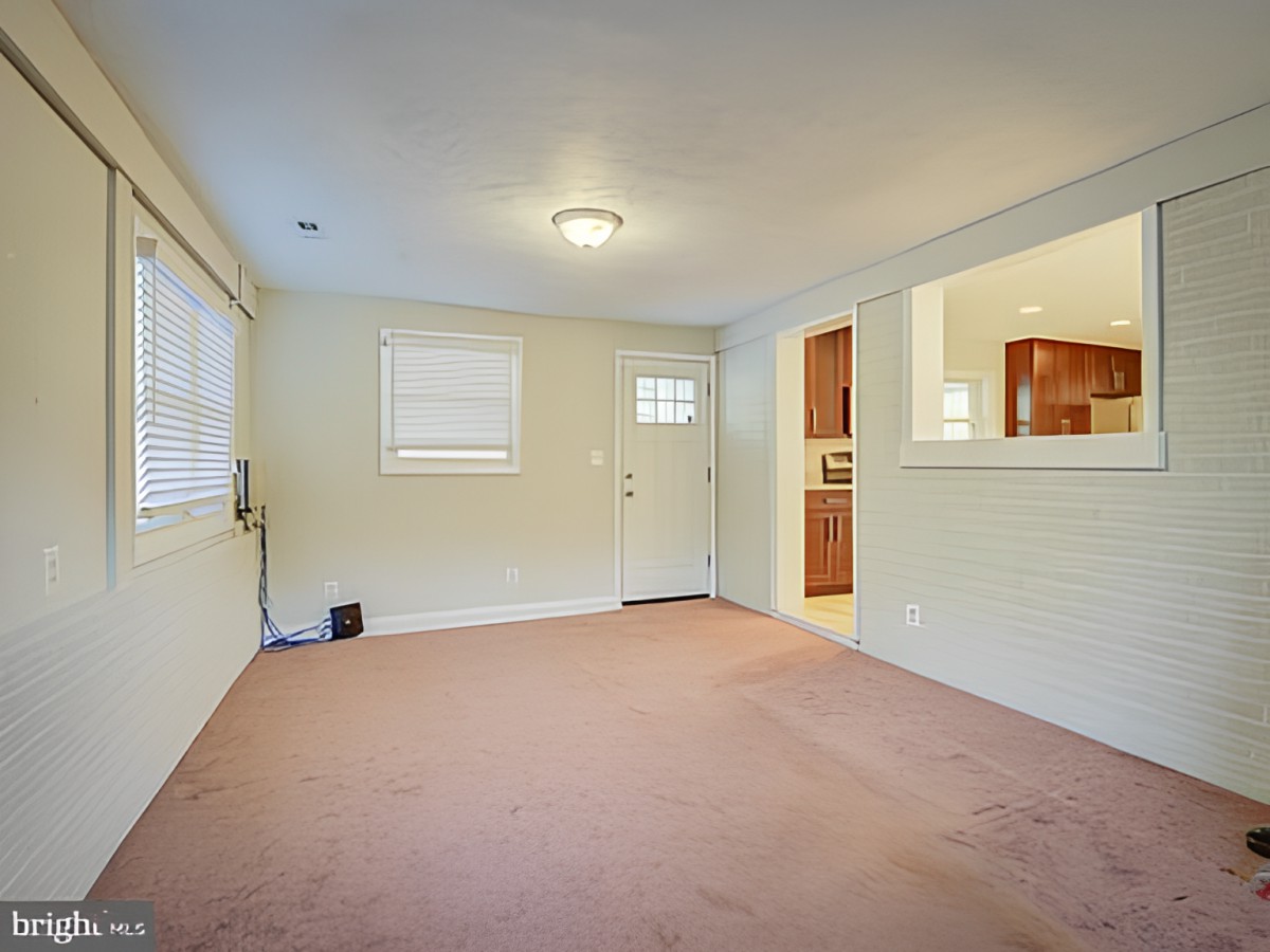 A spacious living room with carpet flooring, featuring multiple windows, a doorway, and an open view into the kitchen area.