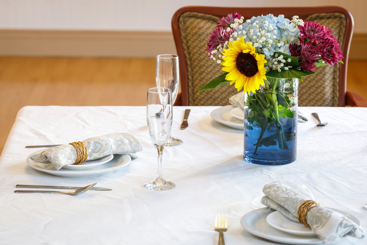A beautifully arranged dining table with elegant glassware, plates, and a vase of colorful flowers including a sunflower centerpiece.