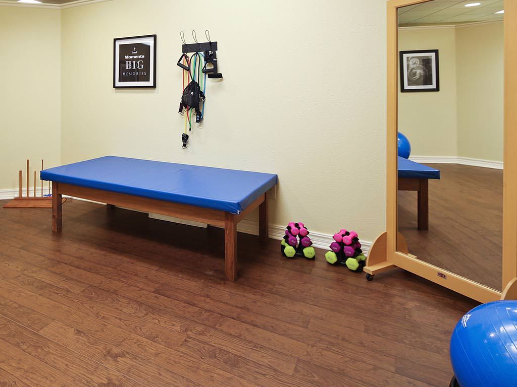 A therapy and exercise room featuring a blue-matted table, a large mirror, and various fitness equipment including dumbbells.