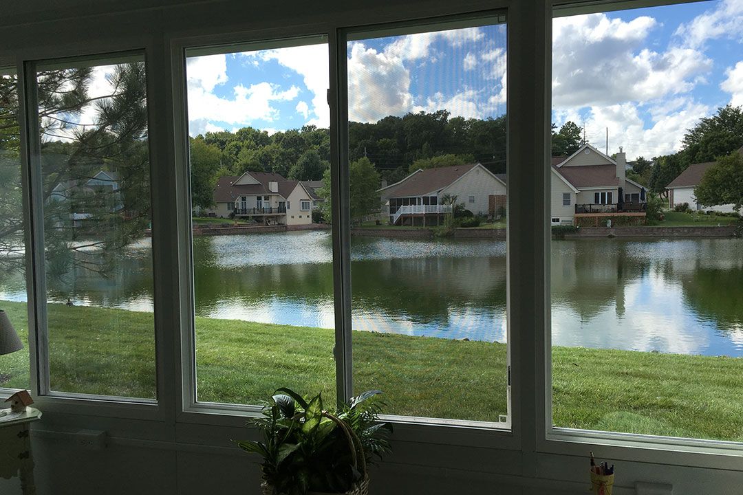 A serene view through a window overlooking a lake with houses and lush greenery under a partly cloudy sky.