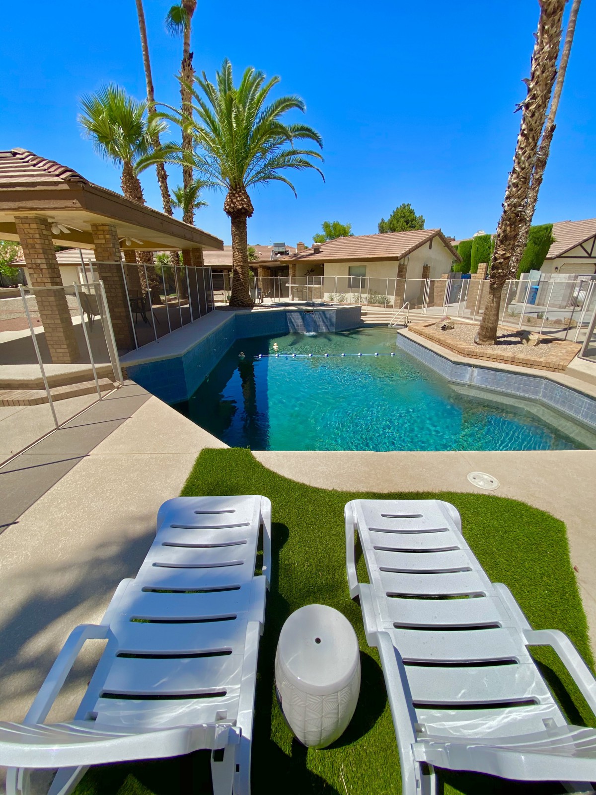 A tranquil residential outdoor swimming pool area with lounge chairs and palm trees under a clear blue sky.