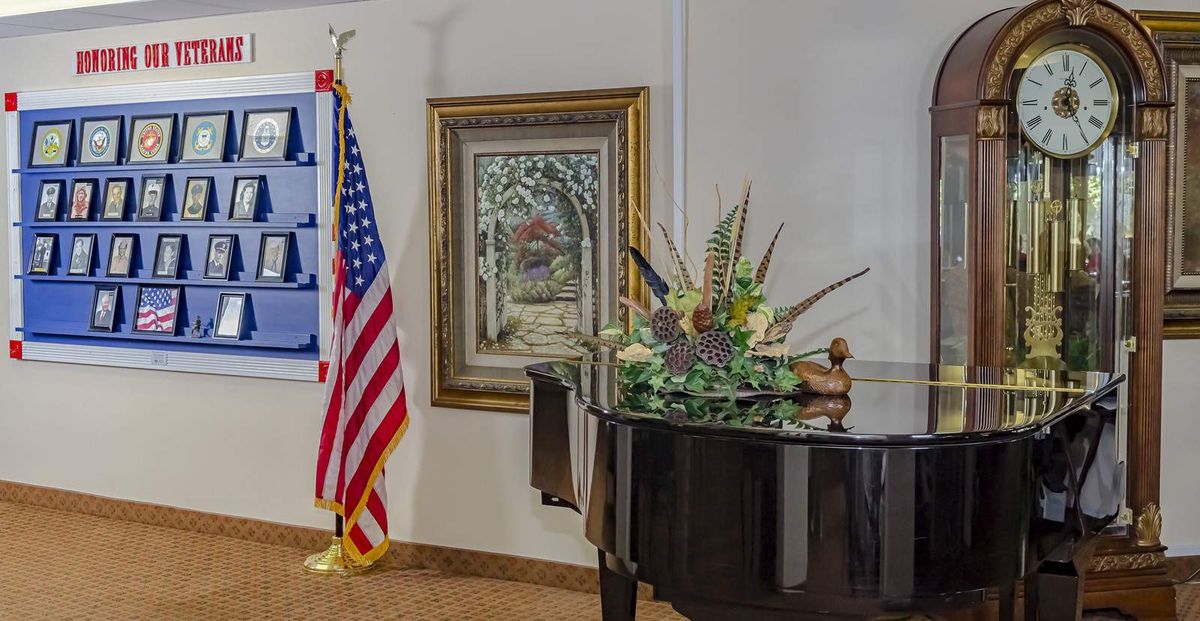 A grand piano decorated with flowers sits in a room featuring an American flag and a wall painting.