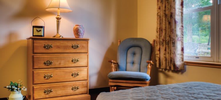 A cozy bedroom setting featuring a blue upholstered chair, wooden dresser, and a lamp providing soft lighting.