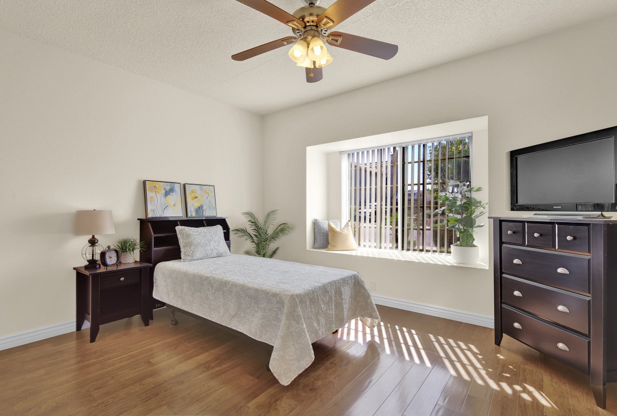A cozy minimalist bedroom with a single bed, wooden flooring, and ample natural light from a large window.