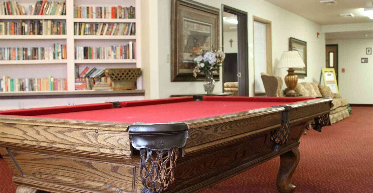 A cozy room features a red-felt pool table, surrounded by shelves of books and decorative elements.