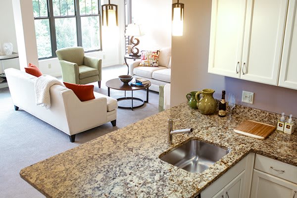 A modern kitchen with a granite countertop overlooks a cozy living room featuring armchairs and a coffee table.