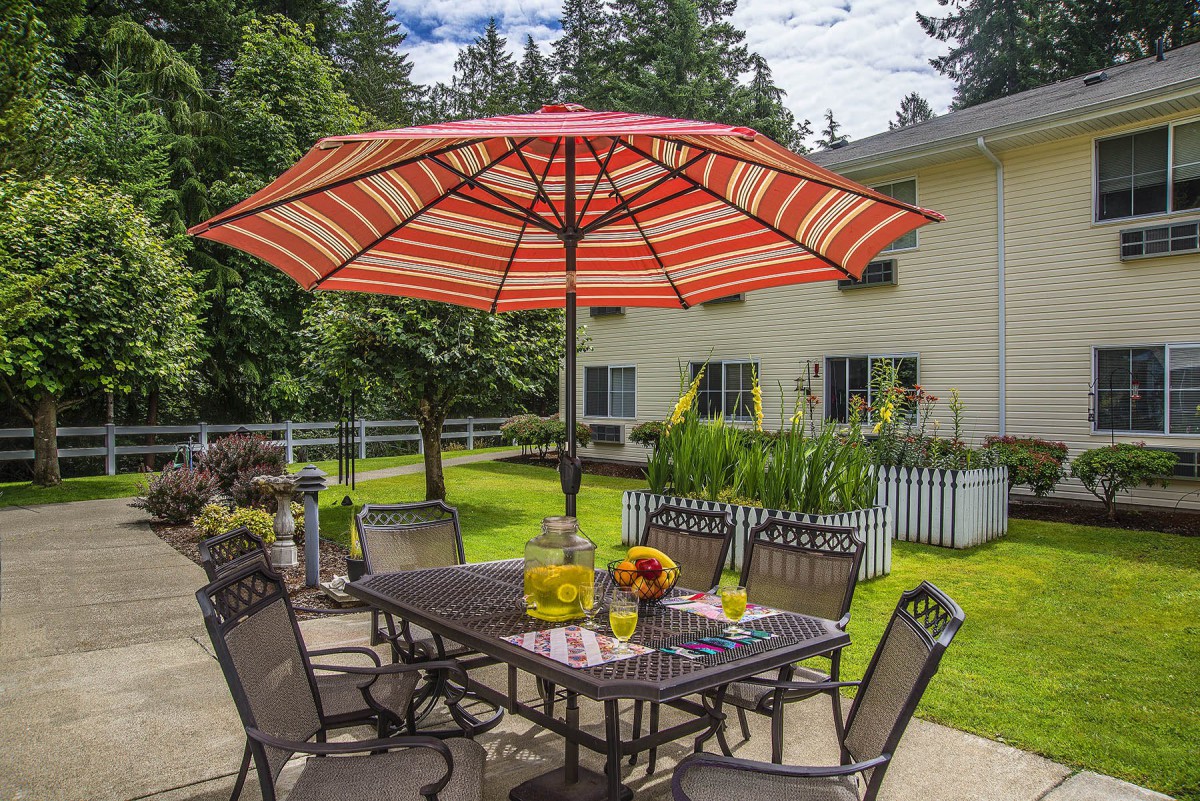 A cozy patio setting with a dining table and chairs under a red-striped umbrella, surrounded by lush greenery.