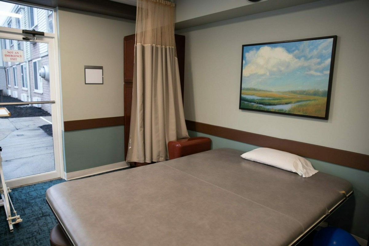 A simple room featuring a bed, pillow, curtain, and a landscape painting on the wall.