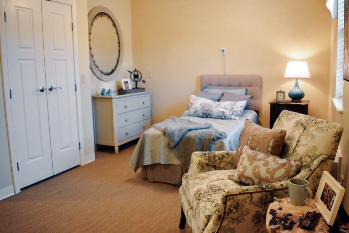 A cozy and elegantly decorated bedroom featuring a single bed, an armchair with floral patterns, a dresser with a mirror above it, and a warm lamp beside the bed.