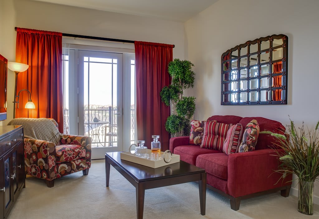 A cozy living room with red accents, featuring a sofa, armchair, coffee table, and large windows with red curtains.