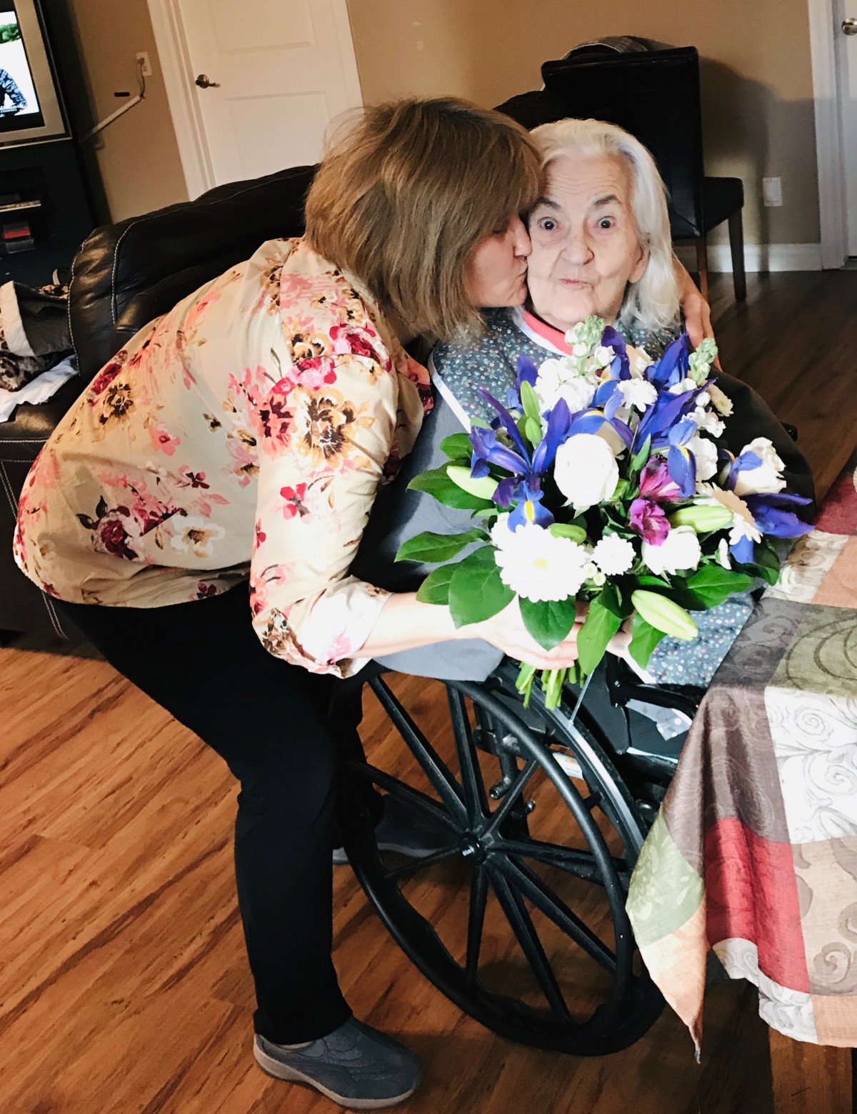 A woman kisses an elderly lady sitting in a wheelchair, holding a colorful bouquet.