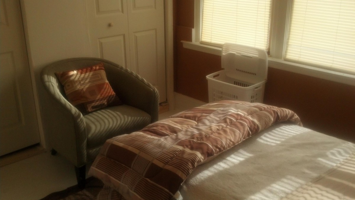 A cozy bedroom featuring a bed, an armchair, and a basket next to a window with blinds.