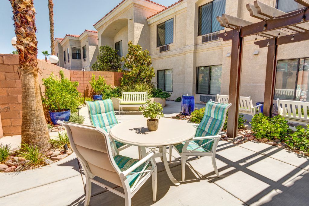 A sunny outdoor patio with a set of chairs and a table surrounded by garden plants and an adjacent building.