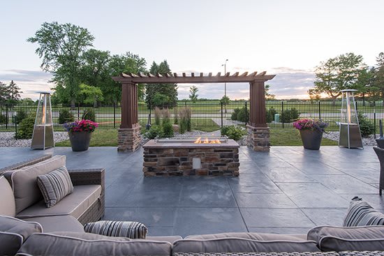 A beautifully designed patio with a fire pit and a pergola, surrounded by lush greenery at sunset.