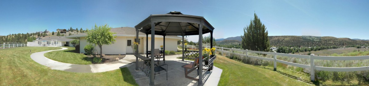 An outdoor gazebo with patio furniture on a sunny day, overlooking a garden and mountains.
