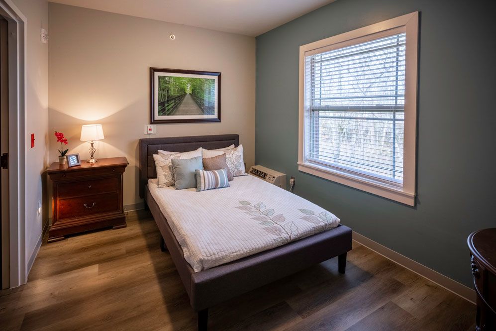 A cozy bedroom with a neatly made bed, nightstand, and window, featuring a soft lamp light and wall art.