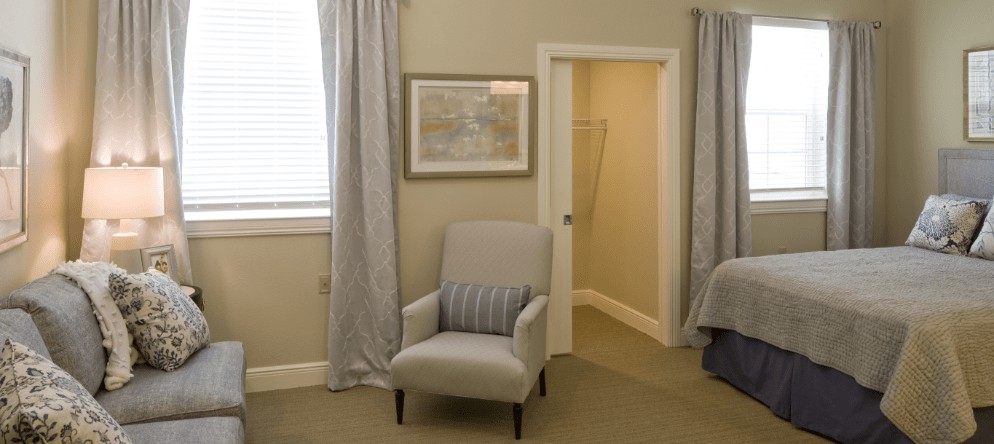 A cozy living room corner featuring an upholstered chair with a striped pillow, framed by long curtains and a wall painting, adjacent to an open doorway.
