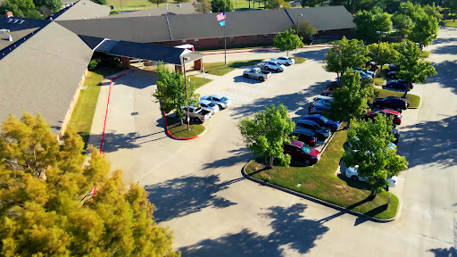 An aerial view of a parking lot filled with cars, framed by lush greenery and a building in the background.