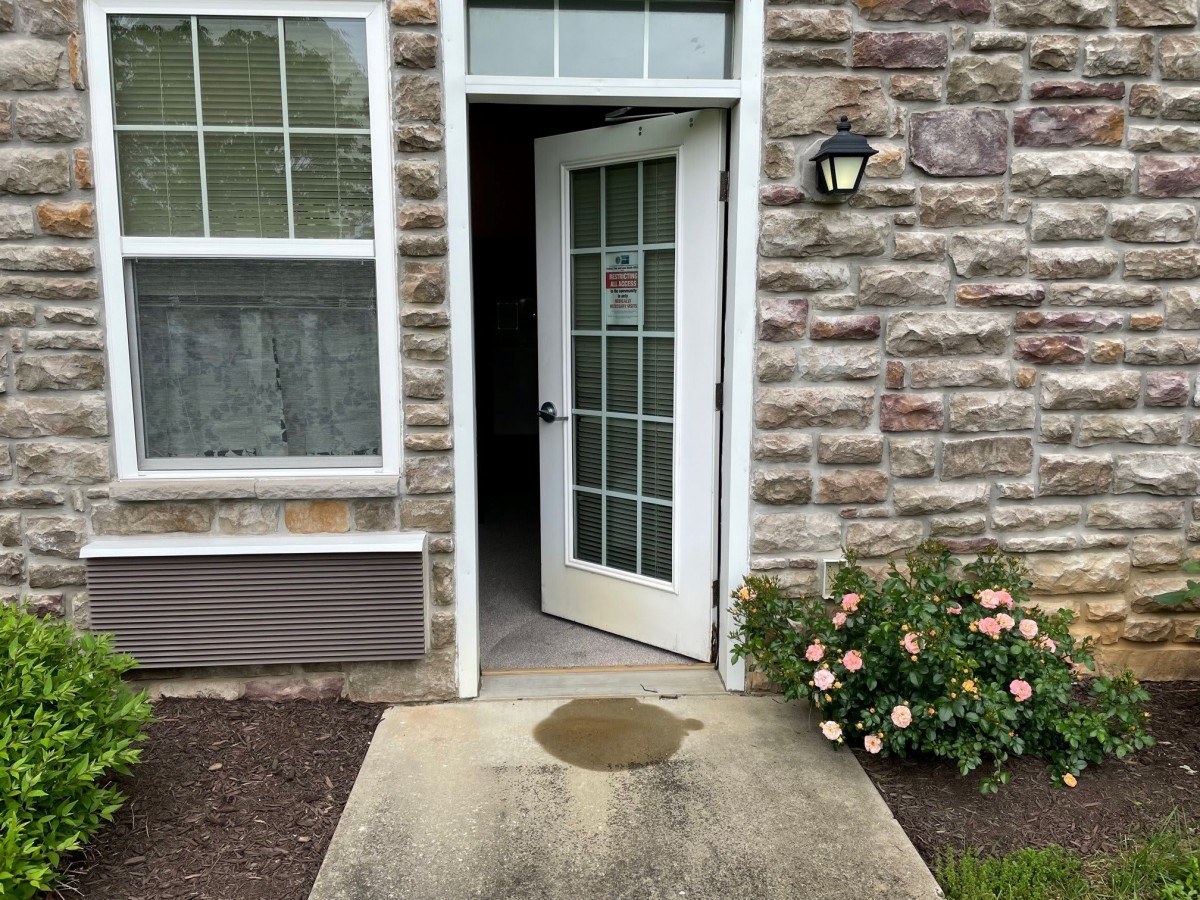 An open white door leading to the interior of a brick building, with flowers and a window nearby.