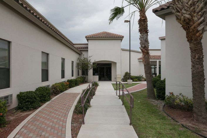 The image shows the exterior of a building with a pathway leading to the entrance, flanked by landscaped greenery and a palm tree.