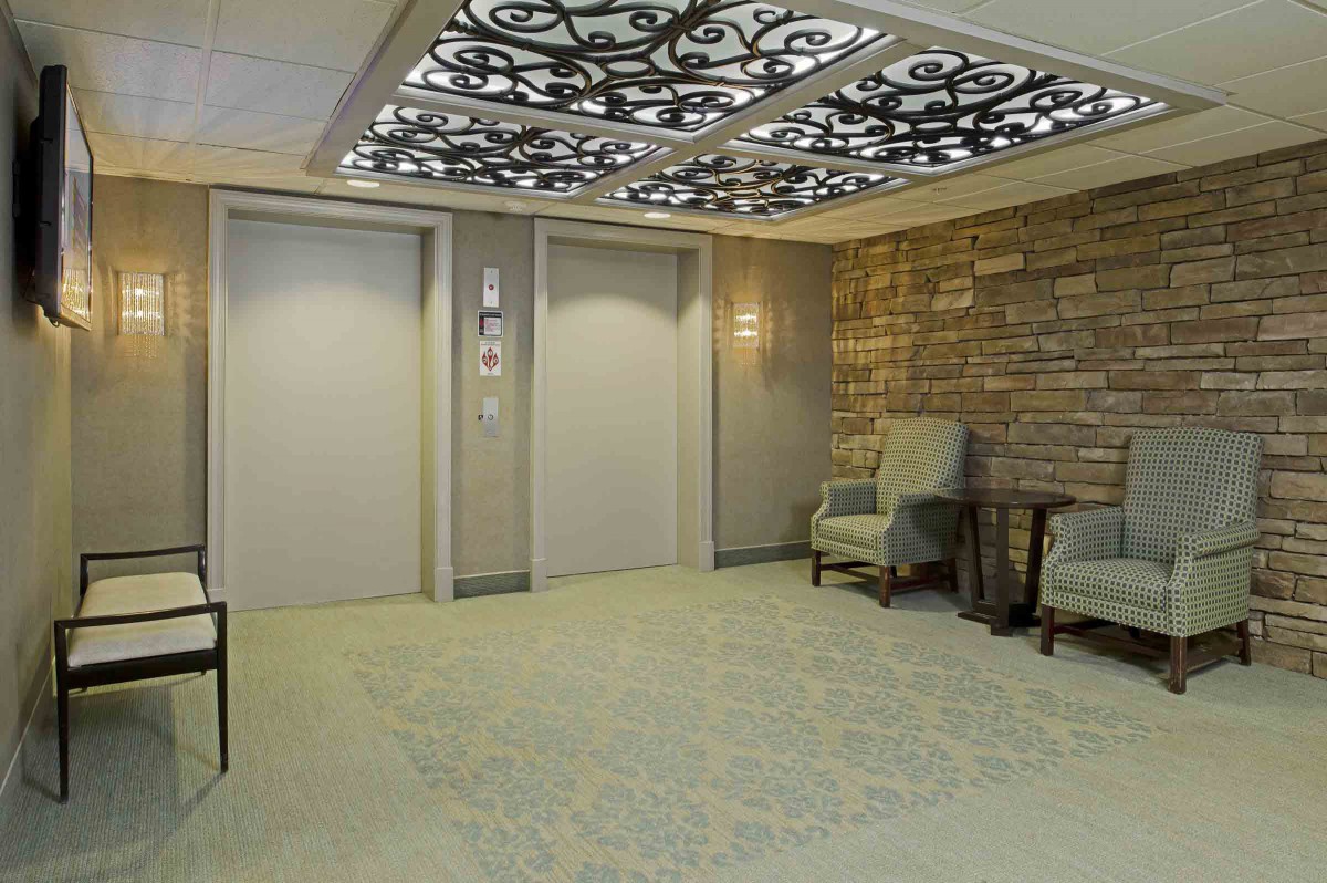A modern elevator lobby featuring decorative ceilings, upholstered chairs, and a stone accent wall.