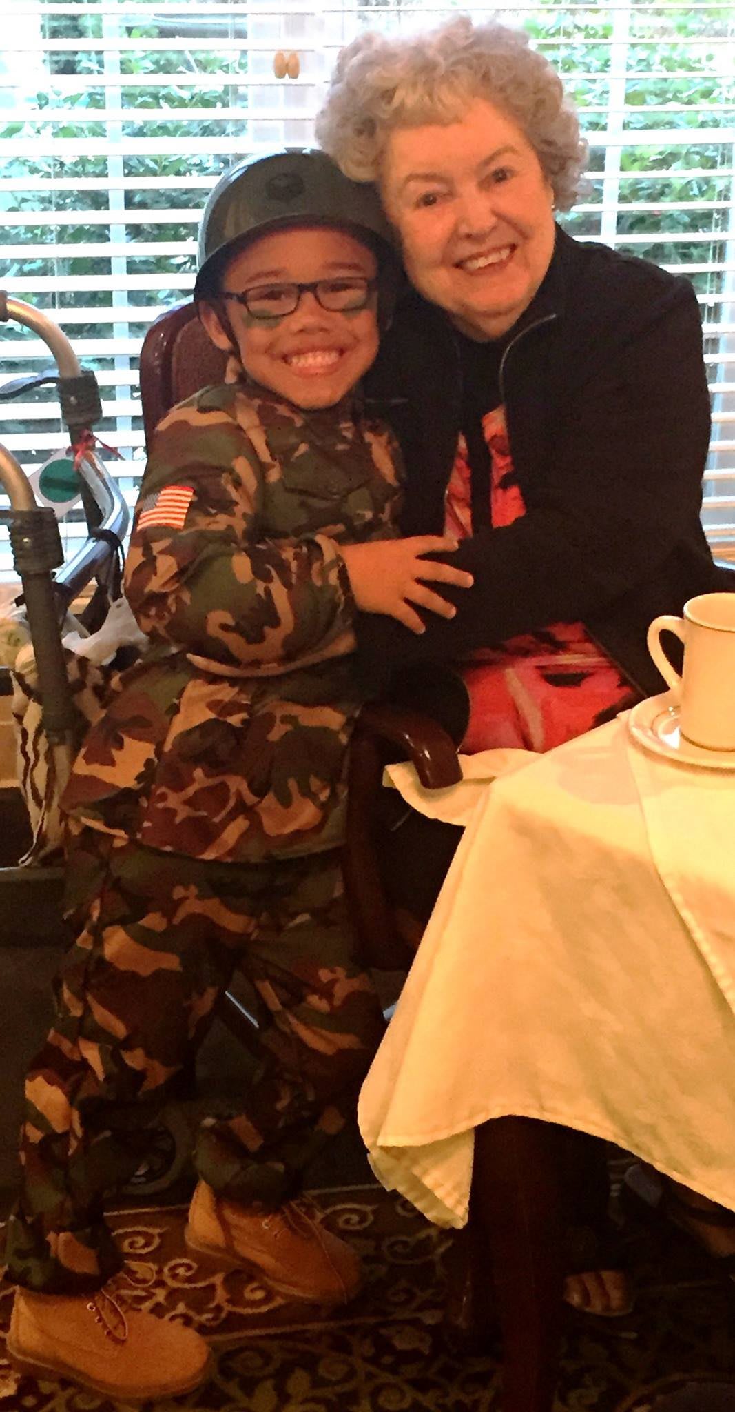 A child wearing a camo outfit smiles while hugging someone at a table indoors.