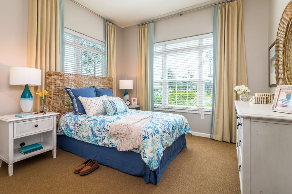 A cozy bedroom with large windows, featuring a bed adorned with blue and floral bedding and surrounded by light-colored curtains.