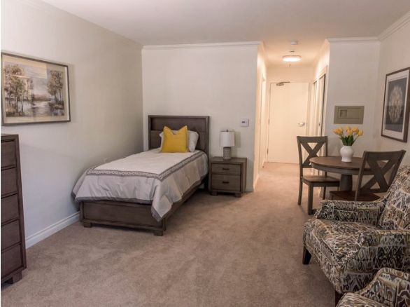 A cozy bedroom with a single bed, nightstand, lamp, and a small dining area.
