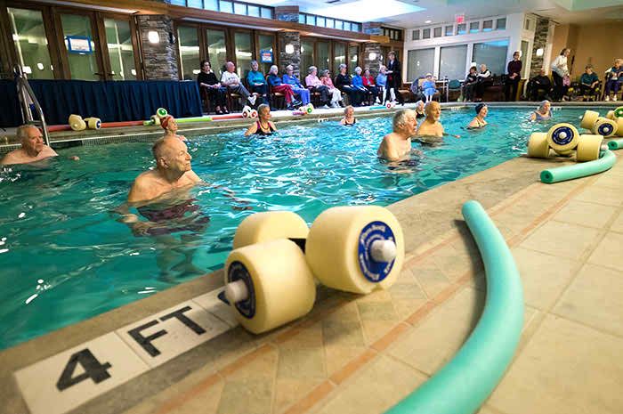 Seniors participating in a water aerobics class at an indoor pool, using foam dumbbells and noodles for exercise.