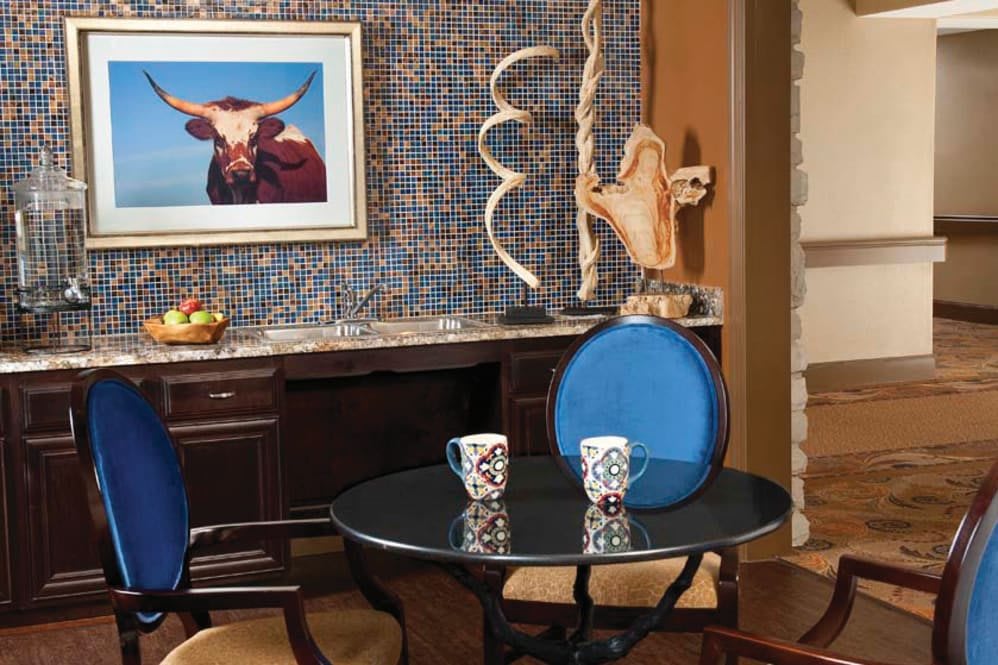 A cozy interior space featuring a small round table with two patterned mugs, surrounded by chairs. The background showcases decorative art and sculptures.
