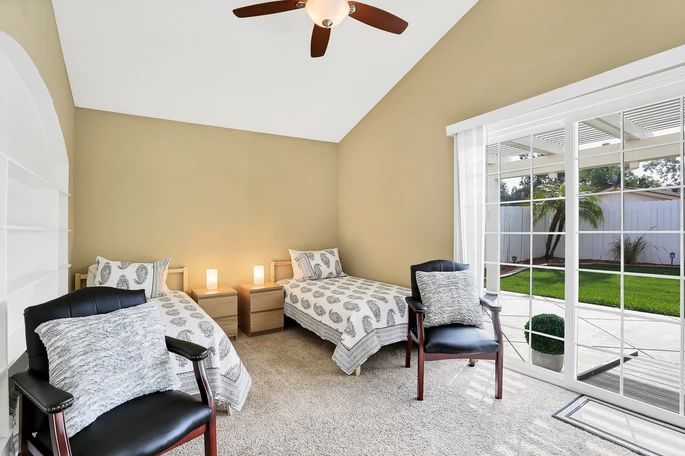 A cozy bedroom with twin beds, carpeted flooring, and a ceiling fan features natural light streaming in through a sliding door.