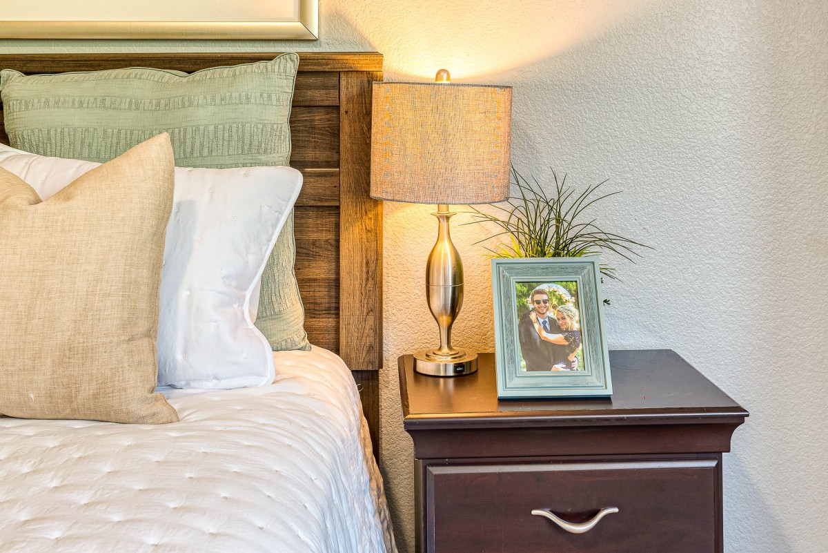 A cozy bedroom scene featuring a nightstand with a lamp, framed picture, and small plant.