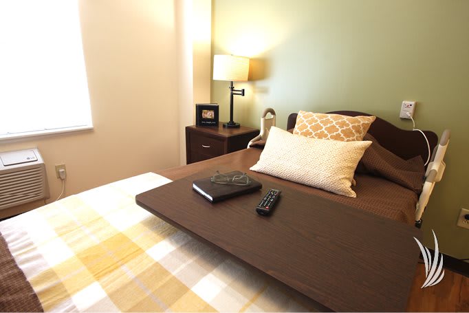 A cozy bedroom with a neatly made bed, featuring a lamp, remote control, and notebook.