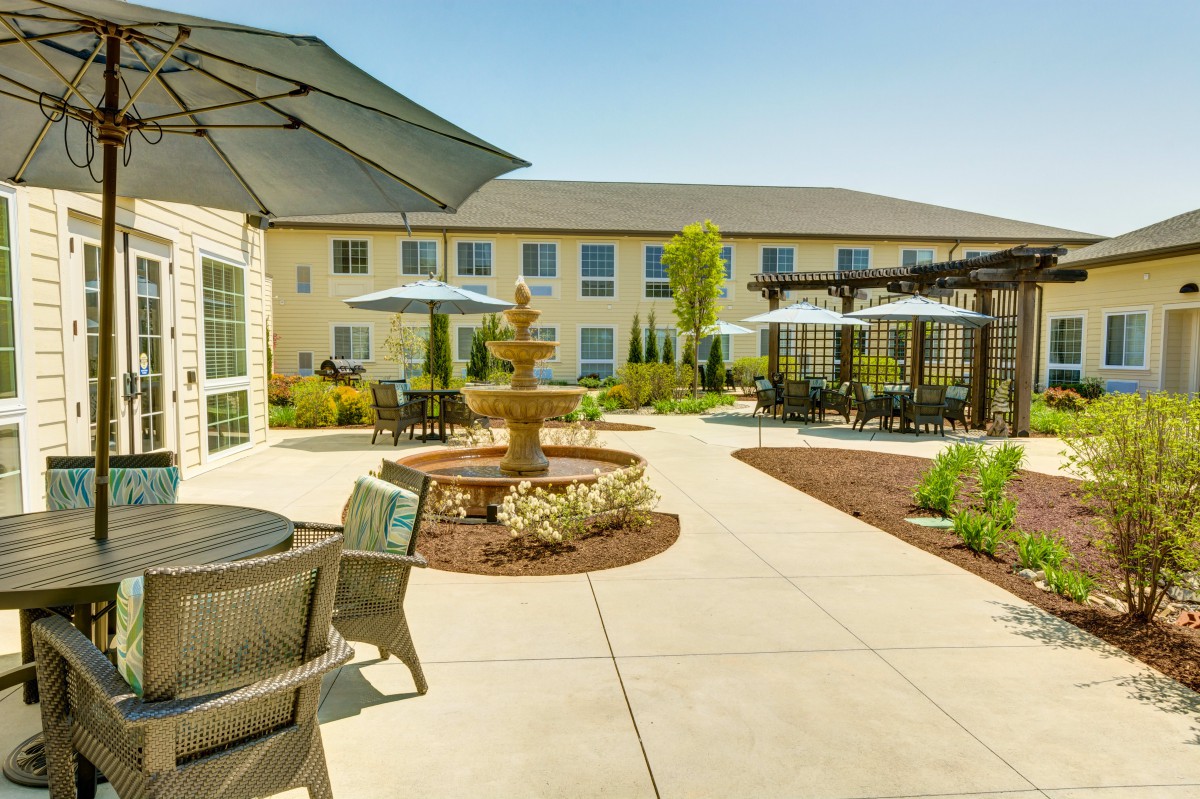 A sunny courtyard featuring a fountain, patio seating with umbrellas, and landscaped garden areas.