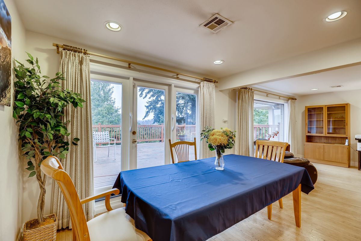A cozy dining room featuring a table with a blue tablecloth, surrounded by wooden chairs and adorned with a vase of flowers. Natural light streams in through the sliding glass doors framed by curtains.