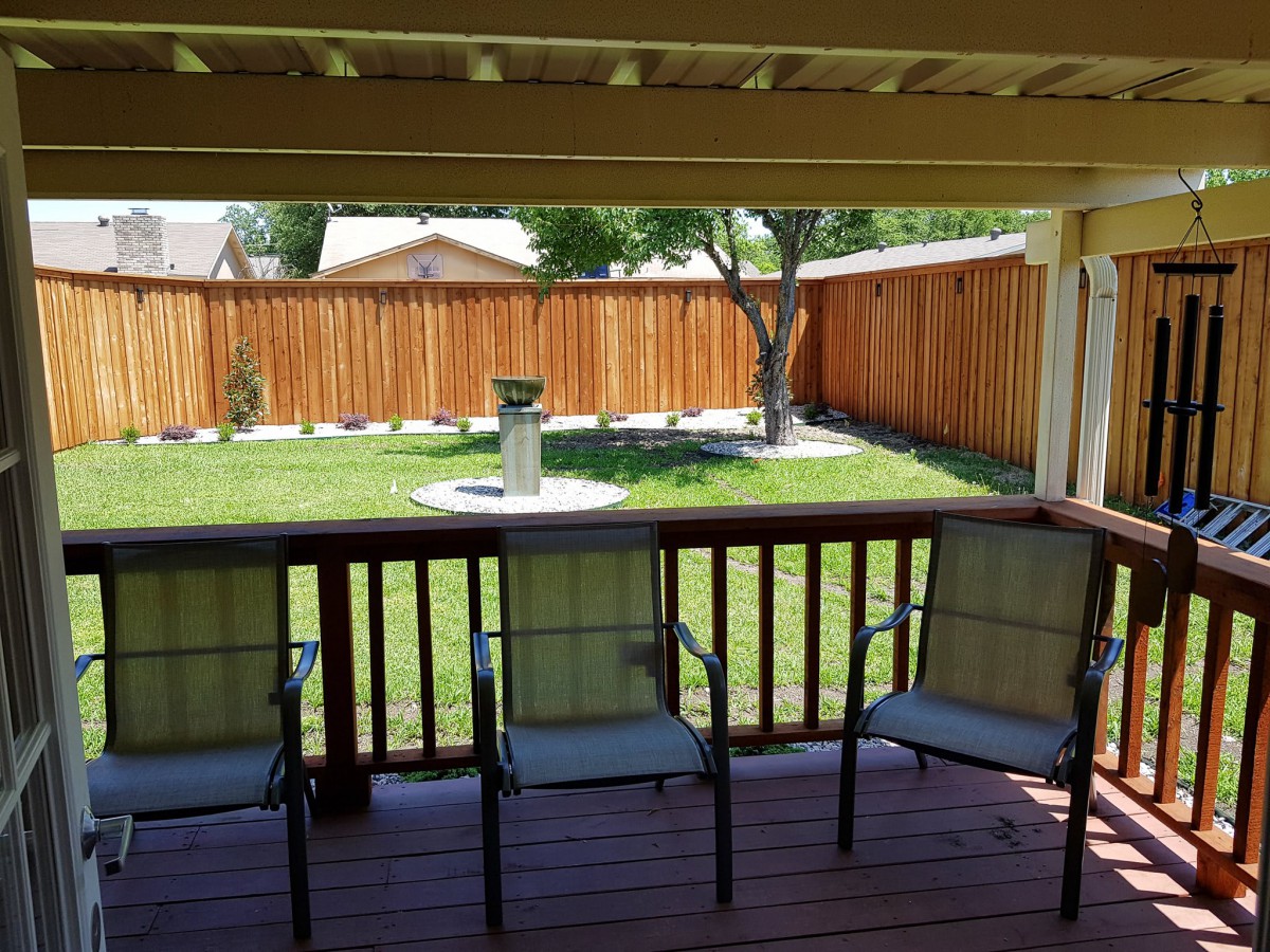 A cozy backyard view from a covered deck with three chairs facing a green lawn and wooden fence.