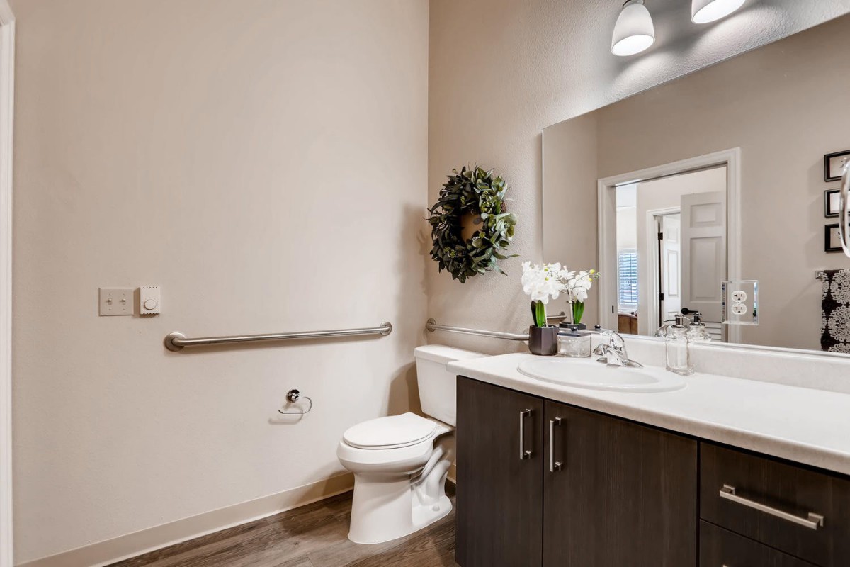 A modern bathroom featuring a toilet, sink with dark cabinetry, and a large mirror accented by a wreath and potted flowers.