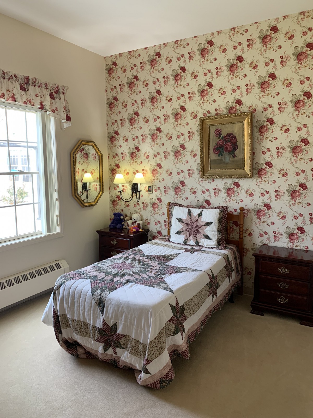 A cozy bedroom with floral wallpaper and a single bed adorned with a quilt, featuring traditional decor elements like a framed artwork and mirror.
